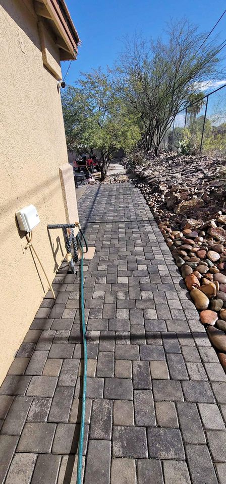 A paved walkway, crafted by expert paver installers using interlocking stone tiles, leads alongside a beige house. A garden hose runs along the path, with desert plants and bushes on the right and a clear blue sky above.