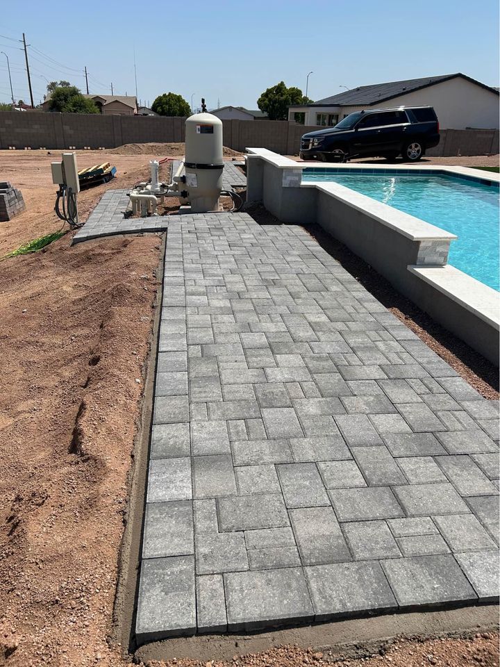 A backyard pool area under construction features a freshly laid stone path, expertly crafted by a skilled paver installer. A filter system stands in the background as landscaping continues, while a parked SUV sits nearby.
