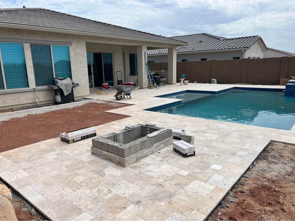 Backyard under construction with pavers installation for a new swimming pool and stone-tiled patio. A rectangular raised structure and several construction materials are on the ground. The house boasts large windows and a covered patio area, perfect for relaxation amidst ongoing improvements.