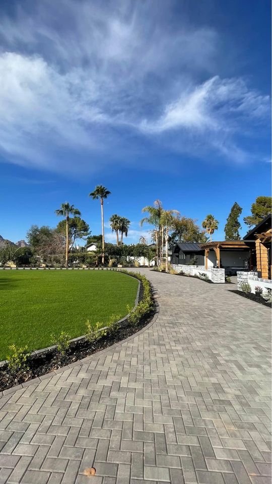 A landscaped garden features a walkway expertly crafted through pavers installation, bordered by green grass and small plants. Tall palm trees stand against a clear blue sky with wispy clouds. To the right, a wooden building is partly visible.