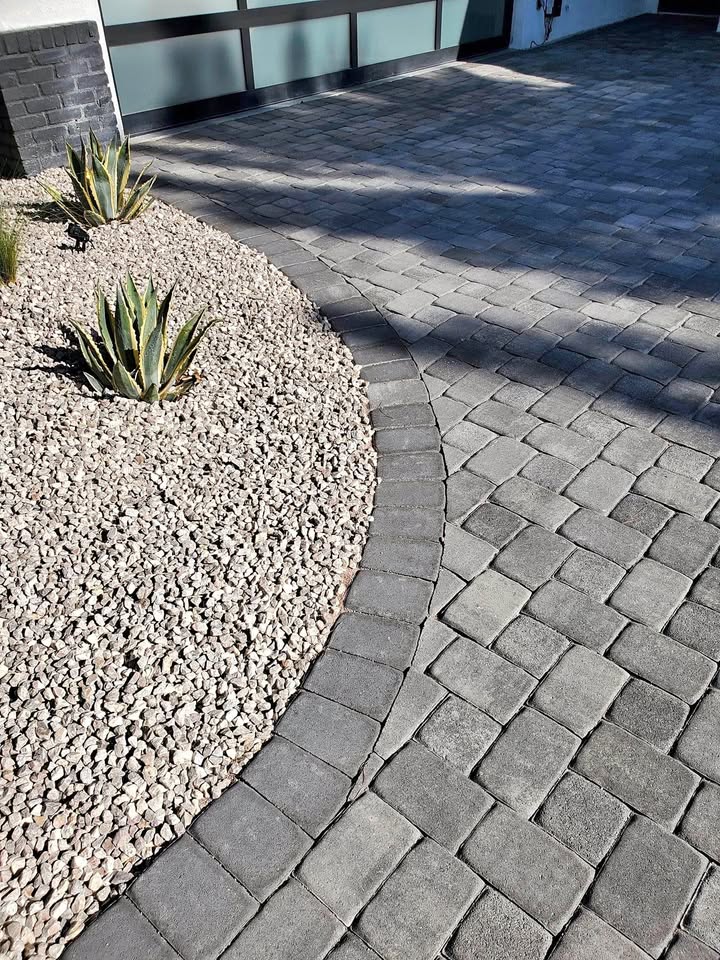 A driveway with expertly installed interlocking gray pavers curves alongside a landscaped area filled with small pebbles and agave plants.