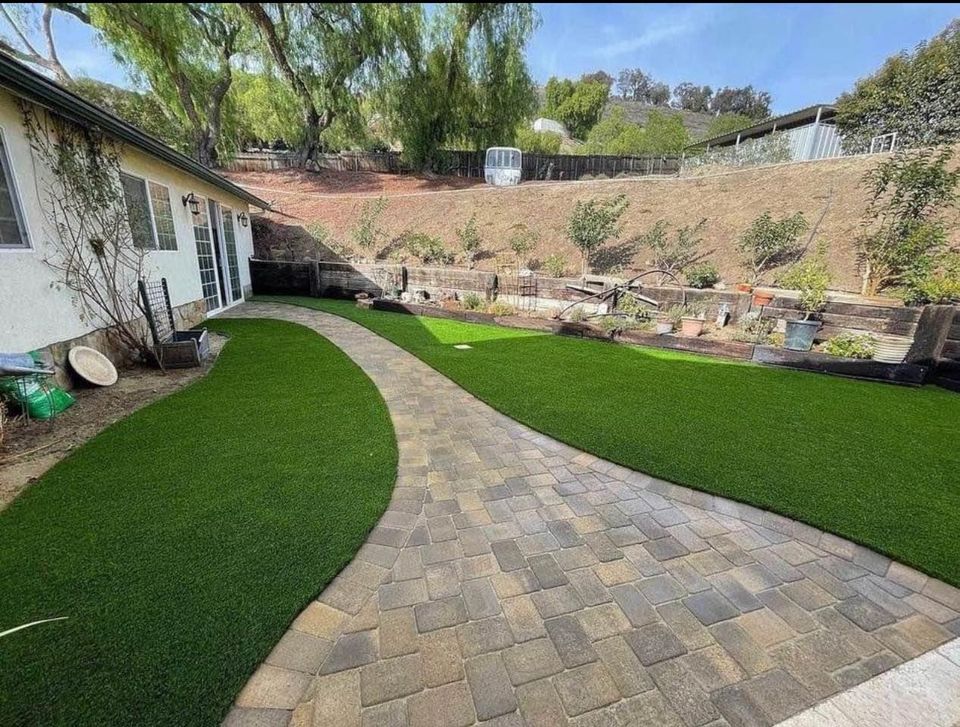 A backyard scene featuring a paved stone pathway, expertly done by a paver installer, curving through lush artificial grass. To the left sits a white house with visible windows and a door. The right side shows a garden with plants and trees, set against a hilly backdrop.