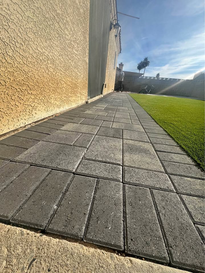 A paved walkway, crafted with precision by expert paver installers, runs alongside a beige stucco wall with large rectangular stones. It leads to a backyard featuring artificial grass, while a palm tree and part of the house stand out under the clear blue sky.