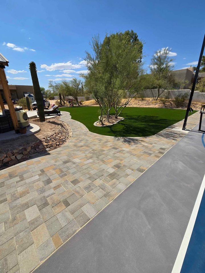 A charming backyard features a tiled patio and lush artificial grass, accented by a few trees. A paved walkway, crafted by expert paver installers, leads to a wooden pergola on the left, while the pool edge gleams in the foreground beneath the clear blue sky.
