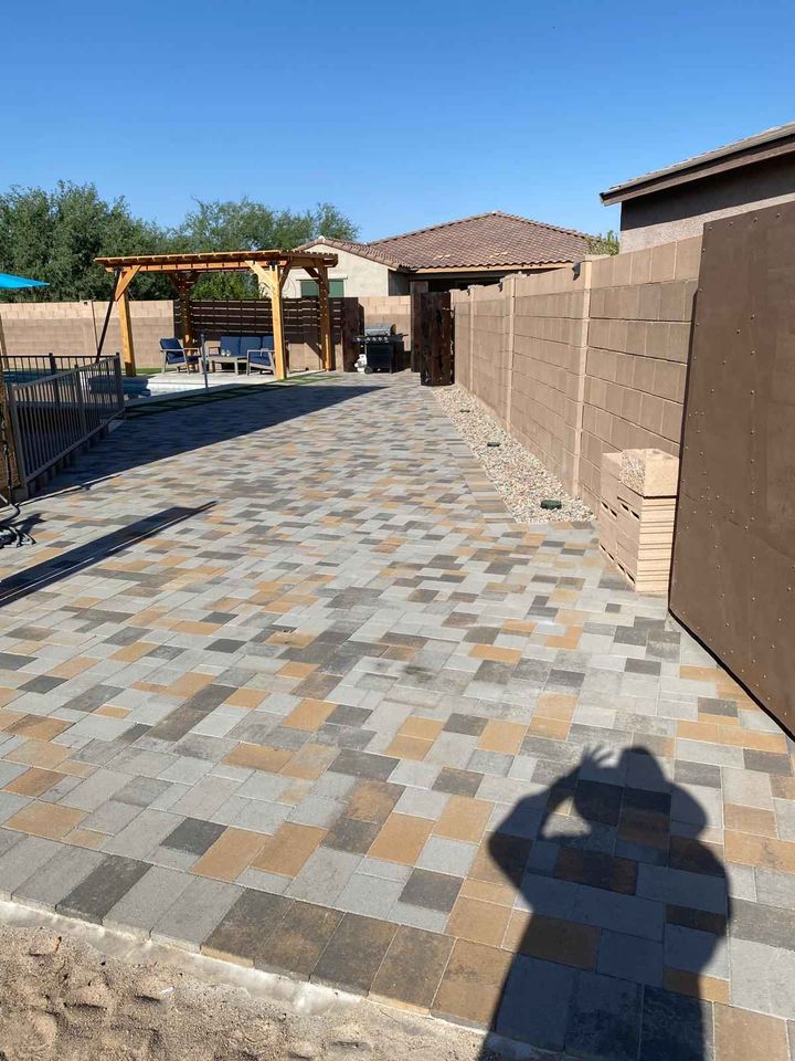 A paved backyard showcases expertly installed gray and tan bricks. A wooden pergola with seating sits invitingly in the distance. Enclosed by a stone wall, the serene space is completed by a person's shadow in the foreground under clear blue skies, a testament to skilled pavers installation.