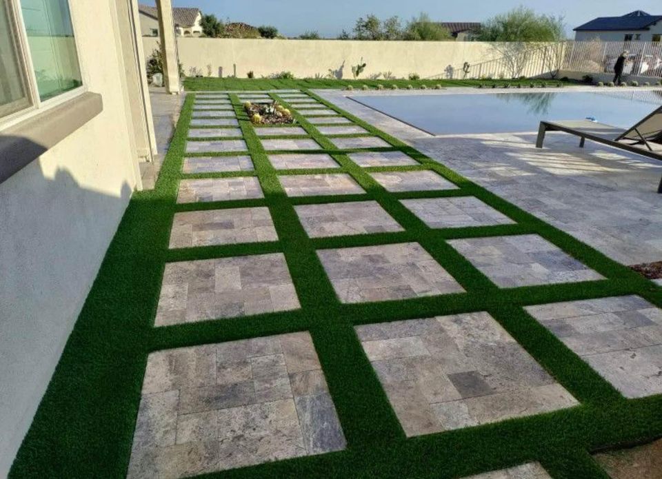A patio with a grid pattern of large stone tiles and strips of lush green artificial grass highlights expert paver installation. This inviting area, adjacent to a swimming pool, features a lounge chair nearby. The surrounding space includes a house and minimal landscaping for a serene retreat.