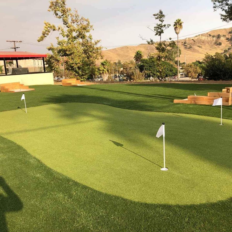 A sunny scene of a backyard putting green with three white flags on poles, surrounded by neatly trimmed grass. In the background, trees stand against distant hills and a building with a red roof.