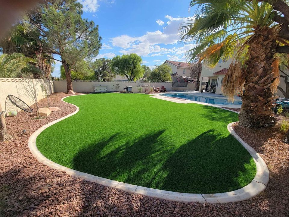 Lush green lawn area featuring backyard putting greens, surrounded by palm and pine trees, adjacent to a pool. The yard is bordered by tan stone walls, with a white two-story house visible in the background under a clear blue sky.