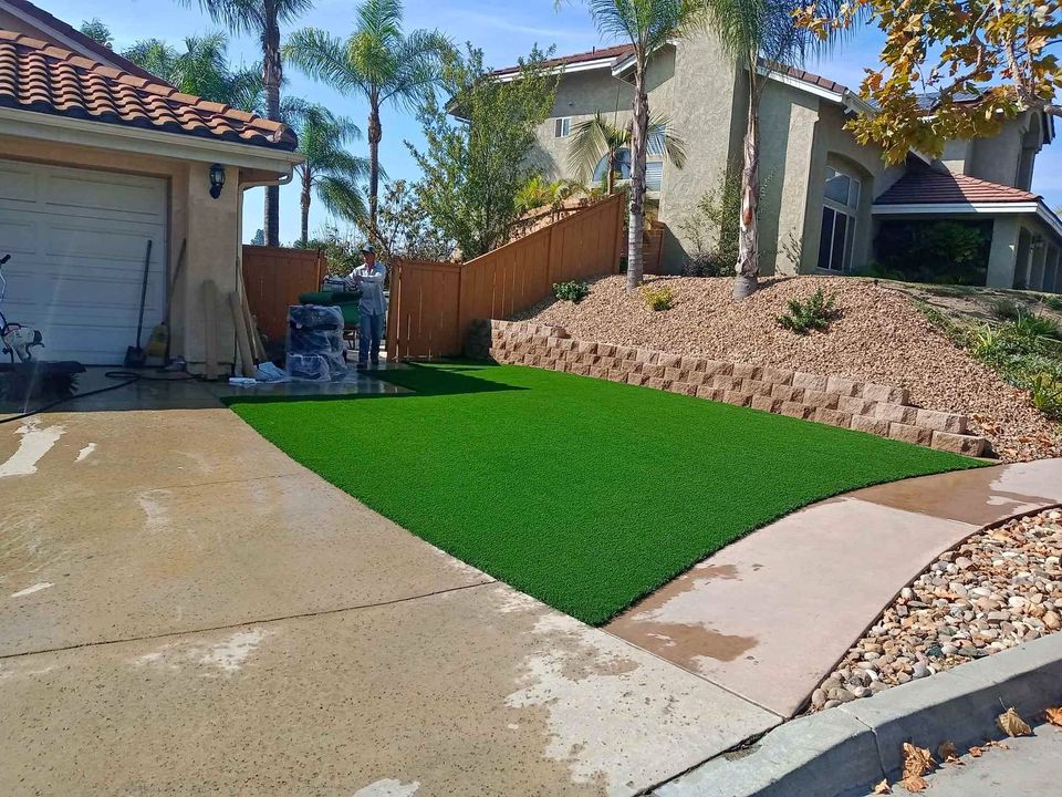 A driveway leads up to a garage with a small patch of artificial turf on the side, perfect for creating backyard putting greens. The house has a beige exterior with palm trees in the background and a landscaped area featuring gravel and a retaining wall.