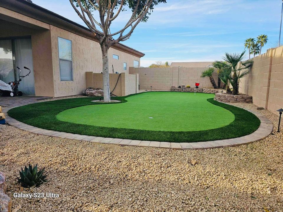 A backyard putting green with a curved design is surrounded by stone tiles, seamlessly blending into the gravel landscape. Desert plants and a stone-framed garden enhance its charm. Perfect for those seeking backyard putting greens, a mower rests by the house wall.