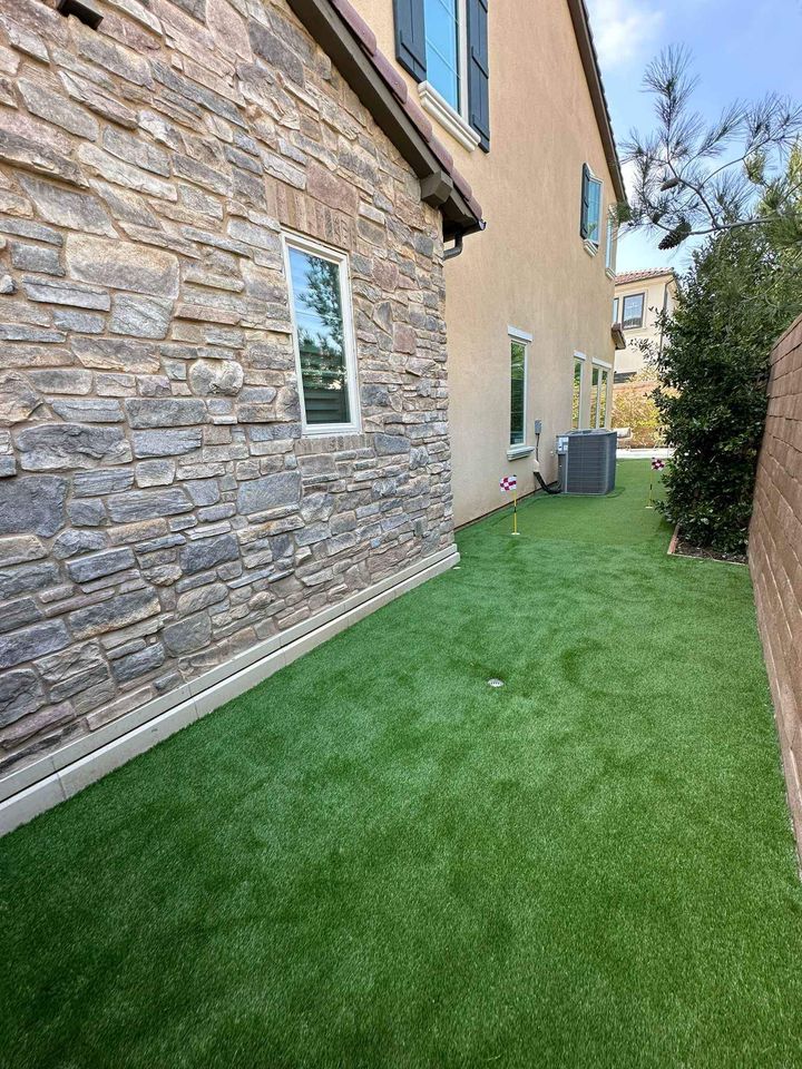 A narrow side yard with artificial grass, perfect for backyard putting greens, runs between two houses. The left house boasts a stone exterior, while the right is partially visible. An air conditioning unit and a small decorative flag accent the serene setting.