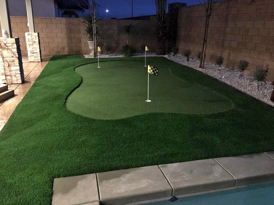A backyard putting green at dusk boasts three golf holes, each marked by yellow flags. The backyard putting greens are surrounded by neatly trimmed grass, a paved area, and the pool's edge. In the backdrop, a stone wall enhances the serene scene with small plants nestled against it.