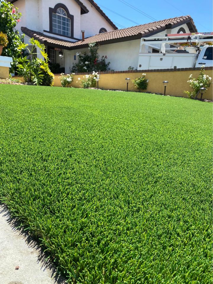 A lush green lawn, ideal for backyard putting greens, leads to a house with a brown-tiled roof and white walls. The lawn is bordered by a yellow low wall with blooming plants and solar lights, while a white vehicle is parked in the driveway.