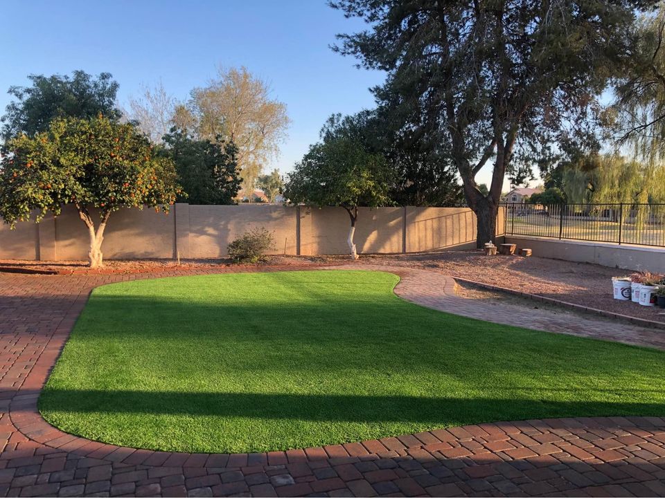 A backyard scene features a green, manicured lawn perfect for backyard putting greens, bordered by red brick pavers. Tall trees and smaller shrubs line the edges. A tan brick wall encloses the area, with a glimpse of neighboring properties visible beyond.