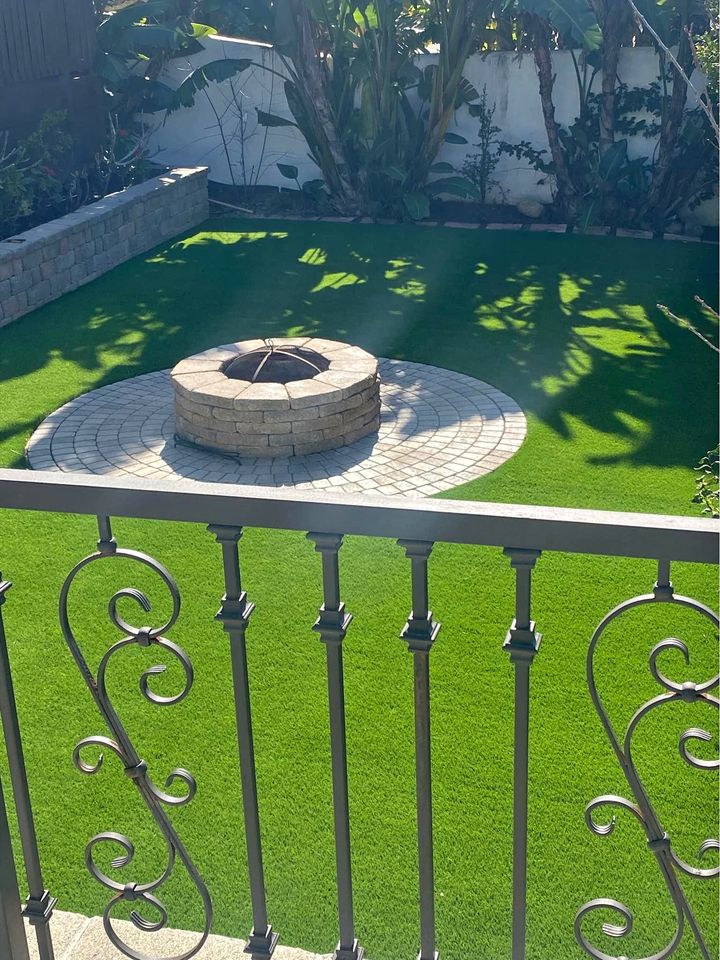 The backyard showcases a circular stone fire pit at the heart of a patio, encircled by lush green artificial grass perfect for backyard putting greens. An ornate metal railing frames the scene, while dense greenery lines the background along a pristine white wall.