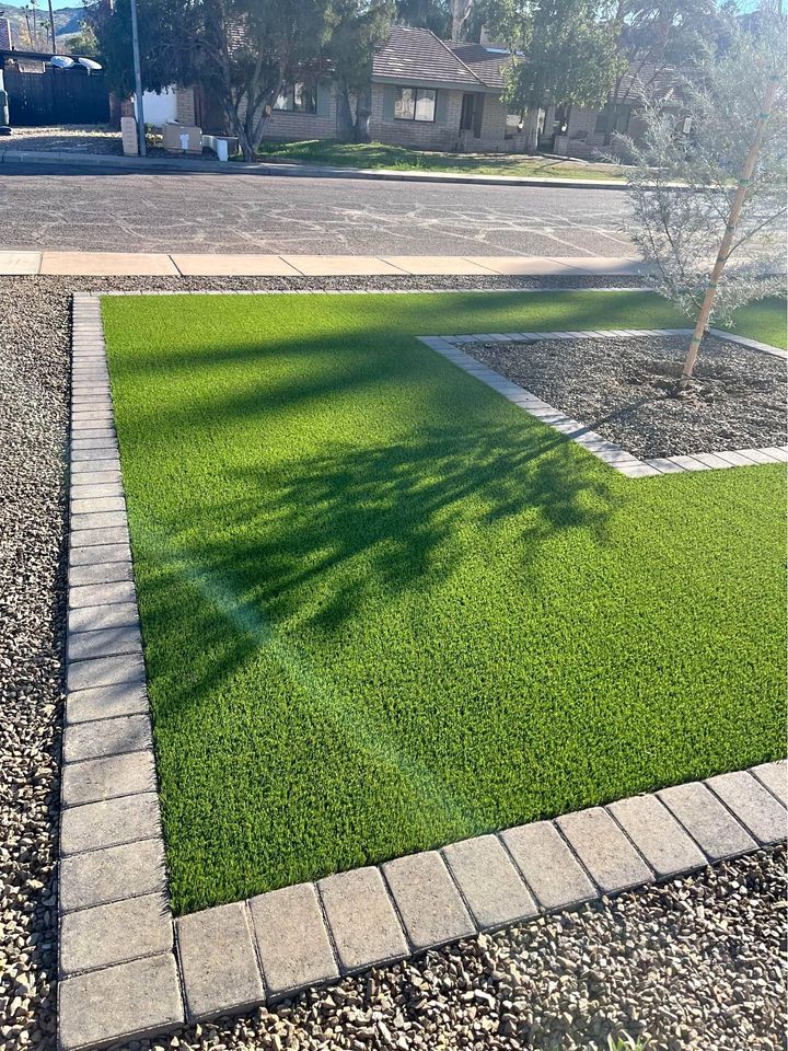 A neatly landscaped front yard features a square patch of vibrant green grass, perfect for backyard putting greens, bordered by rectangular stones. Surrounding the lawn are areas covered with small gray pebbles. In the background, a quiet street with houses and trees is visible.