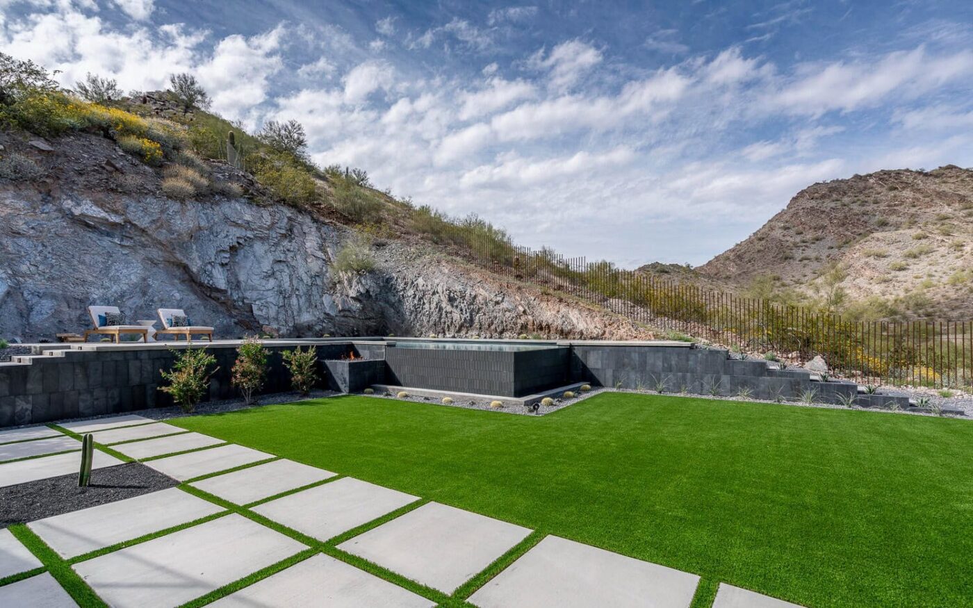 Modern outdoor space with concrete pavers and artificial grass, perfect for backyard putting greens. Surrounded by rocky hills and sparse vegetation, two chairs sit on a raised platform in the background under a blue sky with scattered clouds above.