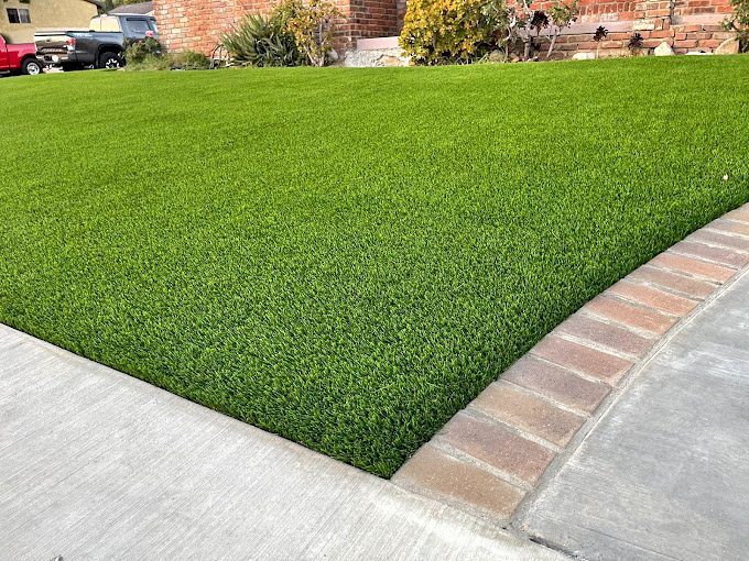 A well-manicured green lawn, reminiscent of backyard putting greens, lies adjacent to a sidewalk and is bordered with brick edging. In the background, part of a brick building and some plants are visible, with a car parked in the driveway.