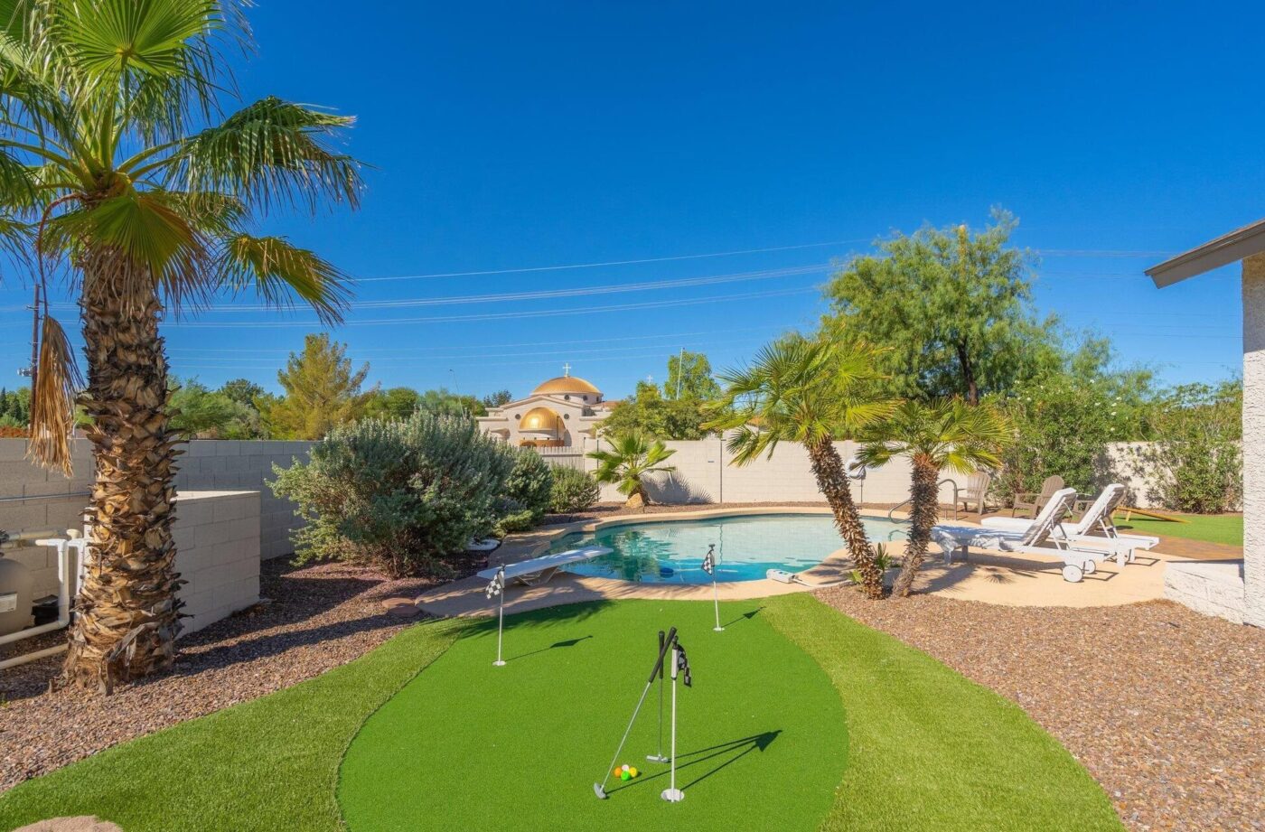 A backyard scene with a small putting green amidst lush tropical plants and a swimming pool. Lounge chairs are perfectly placed for relaxation, while "Backyard Putting Greens" add charm. A blue sky stretches overhead, dotted with bushes and trees in the background.
