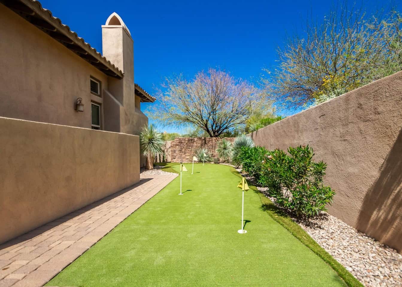 A serene backyard showcasing a rectangular putting green with four hole flags. This ideal space for practicing golf is bordered by desert plants, a stone wall, and a tan stucco house. Under the clear blue sky, a paved walkway complements the charm of backyard putting greens.