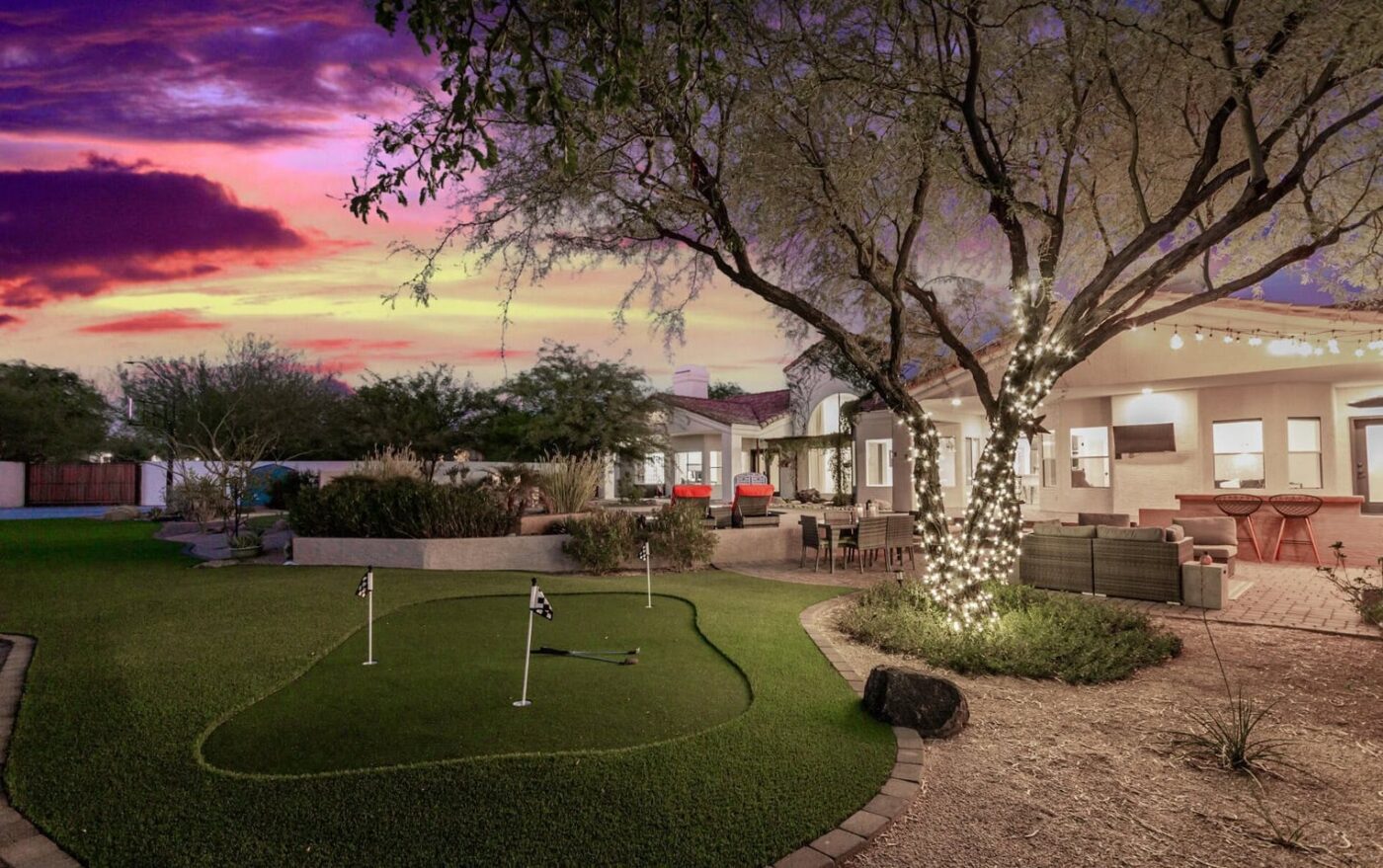 A backyard putting green at sunset is surrounded by lush trees and shrubs. Nearby, a tree wrapped in string lights enhances the modern patio's charm, complete with stylish outdoor furniture. A purple and orange sky casts a warm glow over this serene scene.