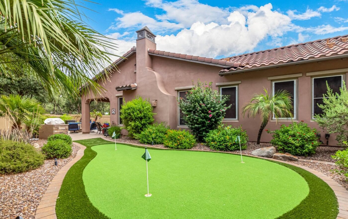 A backyard putting green features three golf holes with flags, surrounded by lush bushes, trees, and a palm. The scene showcases Backyard Putting Greens seamlessly blending with a house's brown stucco walls and tiled roof, all under a partly cloudy sky.