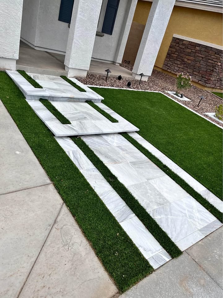 A modern outdoor walkway features white and gray stone slabs arranged in a staggered pattern on a manicured green lawn, expertly crafted by a skilled paver installer. It leads to the entrance of a contemporary house, with white walls and a striking stone accent.
